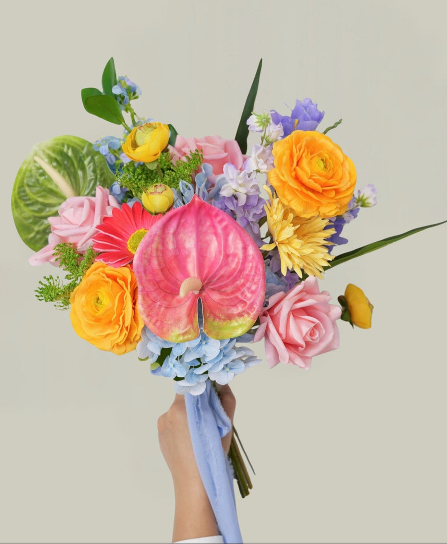 A vibrant, multicolored floral bouquet held by a hand against a solid light beige background. The bouquet features a large pink anthurium, yellow ranunculus, pink roses, blue hydrangeas, purple flowers, and green foliage, tied with a light blue ribbon.
