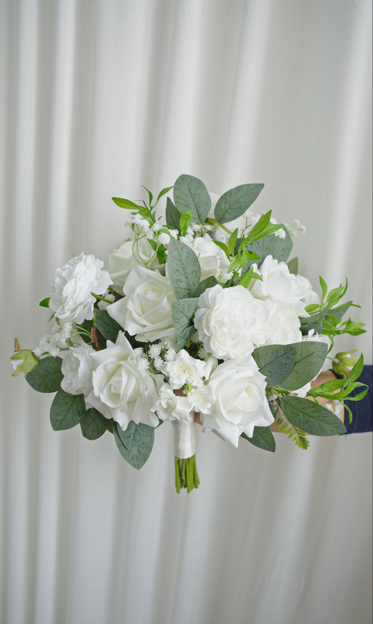 Bouquet of white flowers with green leaves held against a white curtain background