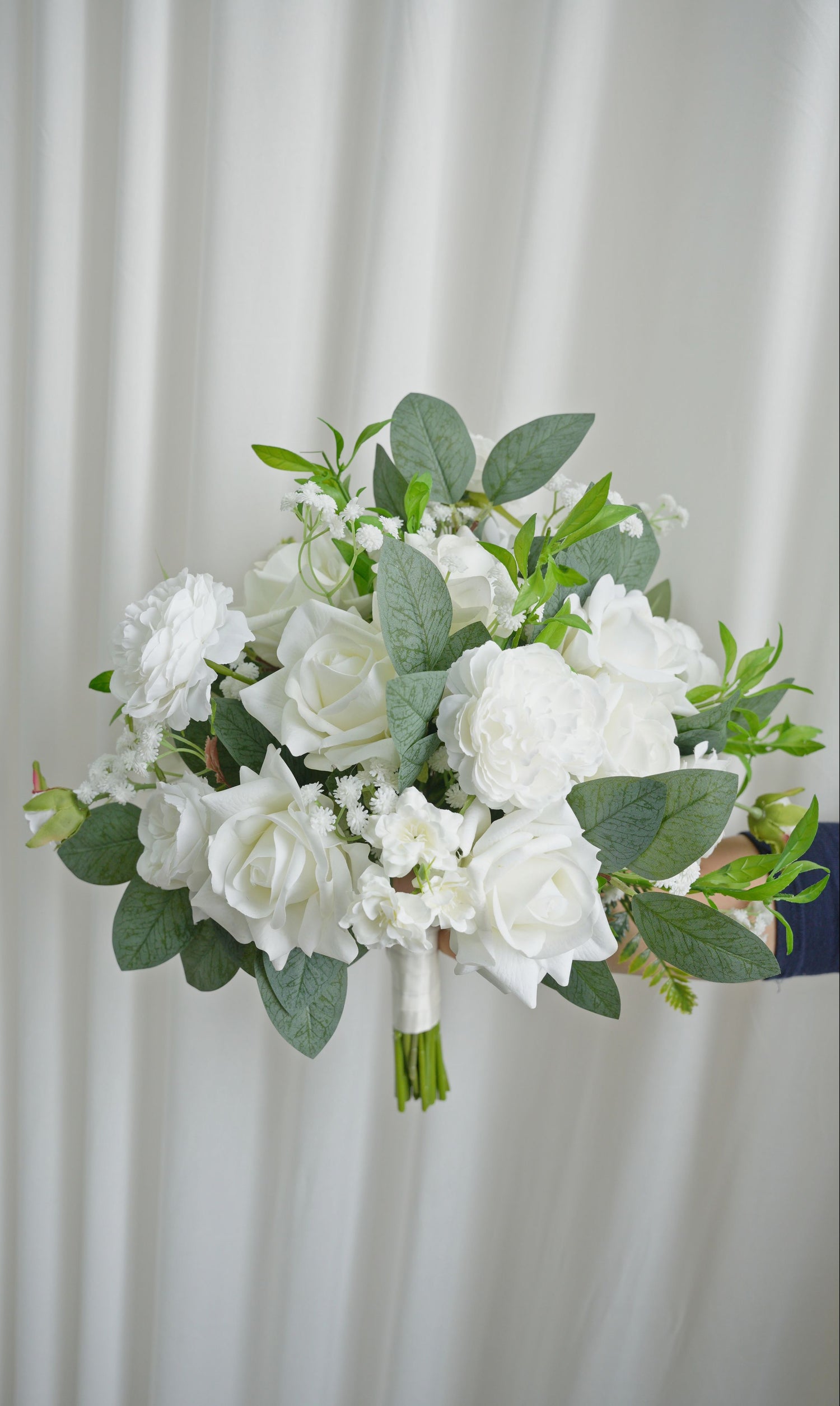 Bouquet of white flowers with green leaves held against a white curtain background
